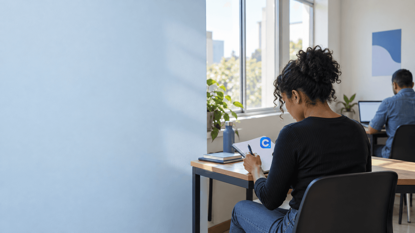 Pessoa anotando em caderno com símbolo Meu Consig em ambiente de trabalho, com outra pessoa ao computador ao fundo.
