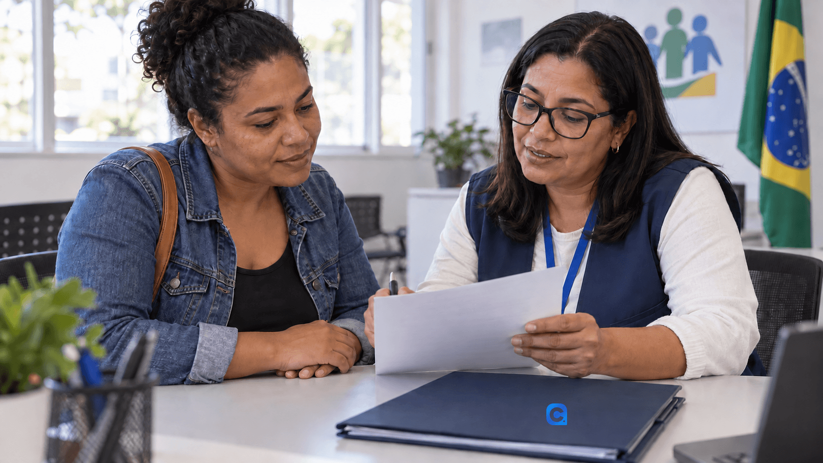 Duas mulheres adultas, uma responsável familiar e uma profissional de atendimento, conversam diante de uma pasta azul em um posto de orientação social; sem crianças ou elementos comerciais.