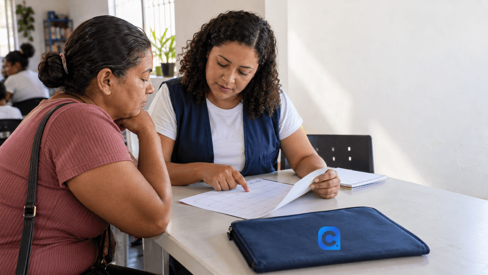 Duas mulheres conferem um calendário sem conteúdo legível em uma sala comunitária; uma pasta azul com o símbolo Meu Consig está sobre a mesa.