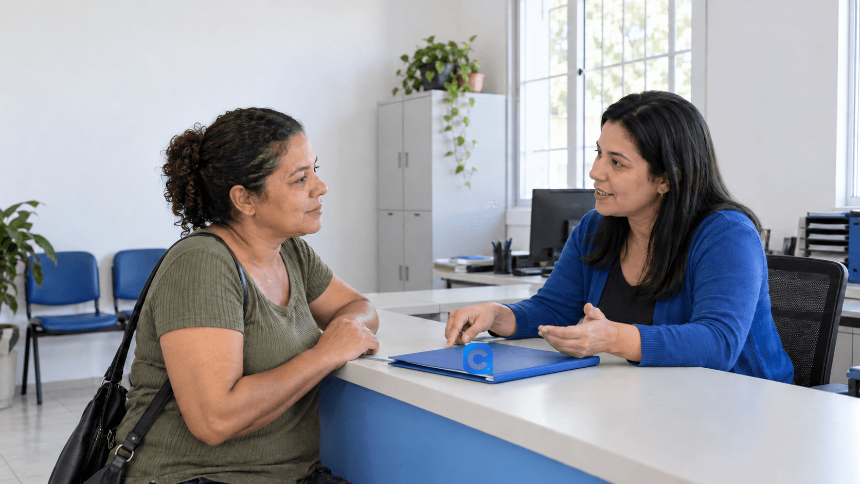 Duas mulheres conversam diante de um balcão de atendimento, com uma pasta azul entre elas.