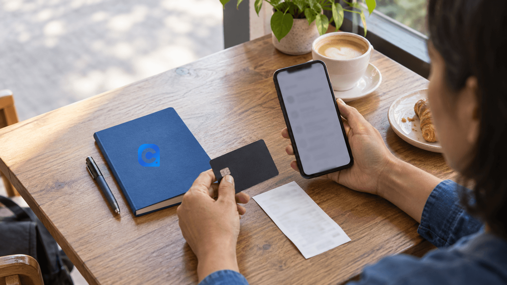 Pessoa adulta confere uma compra no celular enquanto organiza um cartão e comprovantes em um caderno azul com o símbolo Meu Consig.